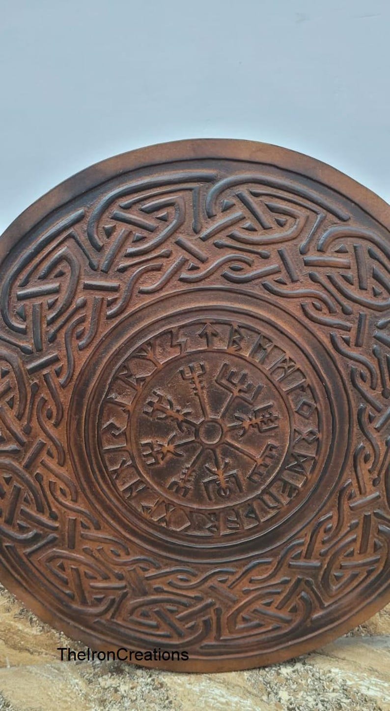 May include: A round, brown, wooden item with intricate Celtic knotwork and a central compass design. The item features a weathered, textured appearance and the text "TheIronCreations" at the bottom. The design is reminiscent of Viking or Norse art.