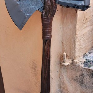 May include: A large, detailed axe with a gray blade and a brown wooden handle. The handle is wrapped with brown leather. The text "TheIronCreations" is visible on the axe.