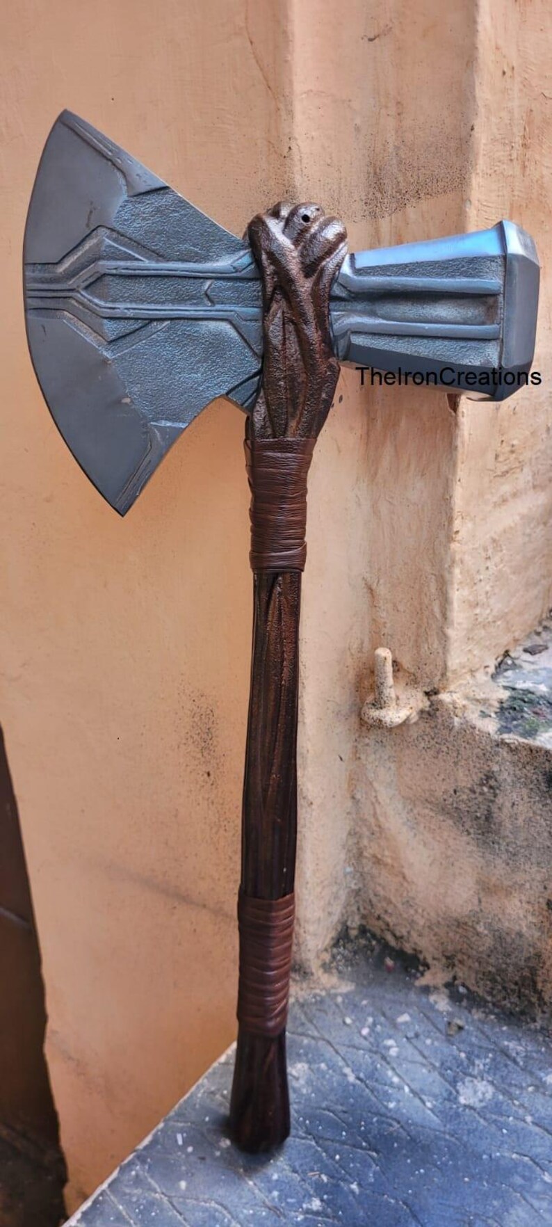 May include: A large, detailed axe with a gray blade and a brown wooden handle. The handle is wrapped with brown leather. The text "TheIronCreations" is visible on the axe.
