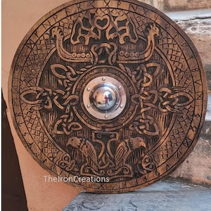 May include: A round, wooden shield with intricate carvings. The shield features Viking ship designs, knotwork, and bird motifs. A central, polished metal boss reflects the surroundings. The text "TheIronCreations" is visible on the lower part of the shield.