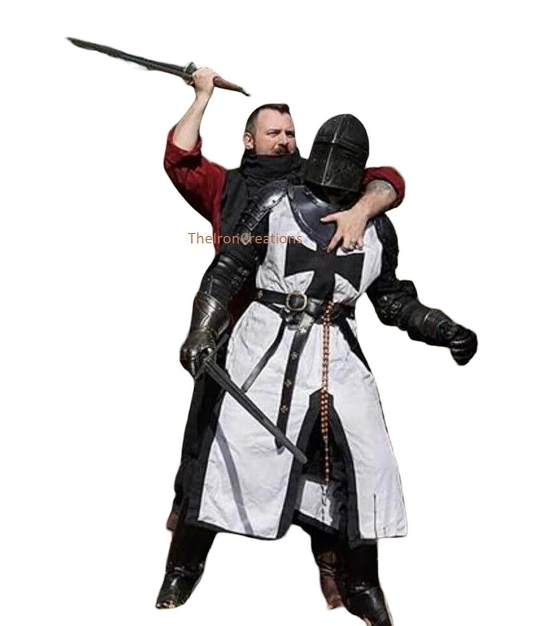 May include: A person in a red shirt and black scarf holds a sword above a person wearing a black and white medieval knight's armor. The armor features a black cross on the chest and a black helmet.