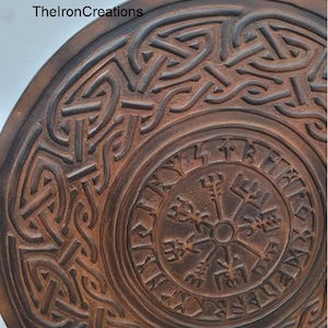 May include: A round, brown, carved wooden object with intricate Celtic knotwork and a central compass design. The text "TheIronCreations" is visible on the upper left. The object has a weathered appearance.
