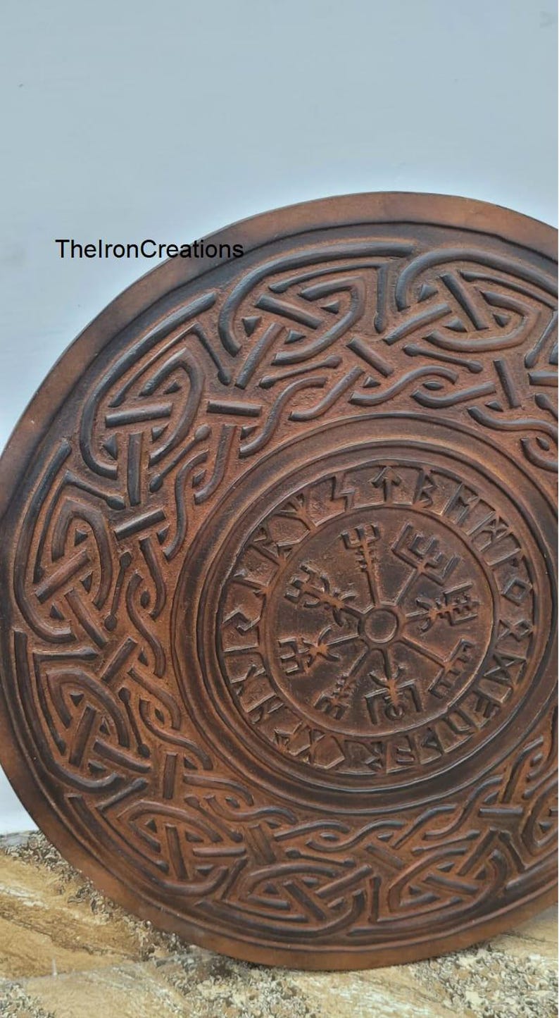 May include: A round, brown, carved wooden object with intricate Celtic knotwork and a central compass design. The text "TheIronCreations" is visible on the upper left. The object has a weathered appearance.