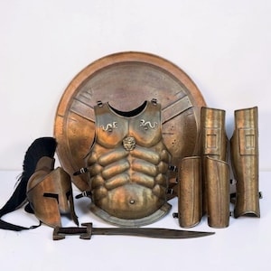 May include: A bronze-toned metal armor set including a circular shield, a muscle cuirass with a central lion head emblem, a helmet with a black crest, a sword, and four pieces of leg and arm greaves. The armor features a weathered, metallic finish.