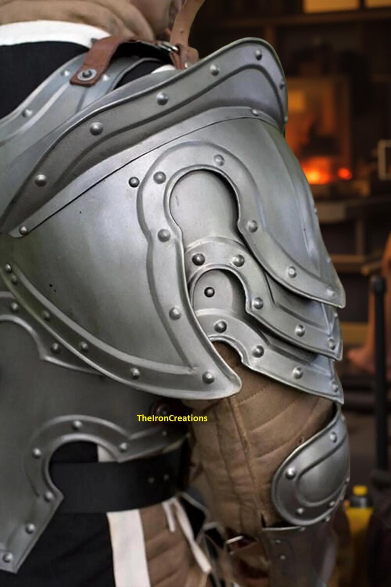 May include: Close-up of a silver-colored metal shoulder armor with riveted details. The armor is layered and curved, with a brown leather strap visible. The text "TheIronCreations" is at the bottom.