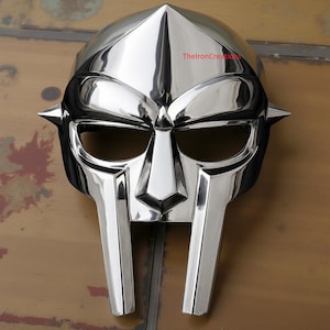 May include: A polished silver mask with a distinctive angular design. The mask features a pointed top, eye openings, and a prominent nose piece. The mask has a reflective surface and is likely made of metal. The text "ThelronCreations" is visible on the top of the mask.