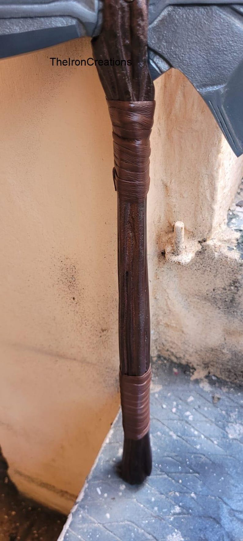 May include: Close-up of a prop axe with a dark brown wooden handle wrapped in brown leather. The axe head is gray. The text "ThelronCreations" is visible on the side.