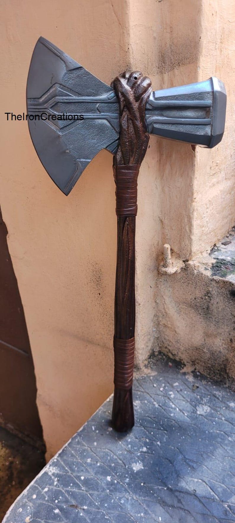 May include: A prop axe with a dark gray blade and a brown handle. The handle is wrapped with brown leather. The axe head has a rectangular shape on one side. The text "ThelronCreations" is visible on the blade.