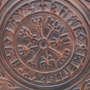 May include: Close-up of a brown, circular object with a raised, intricate design. The center features a compass-like symbol surrounded by runes. The outer edges display a repeating knot pattern. The text "TheIronCreations" is visible.