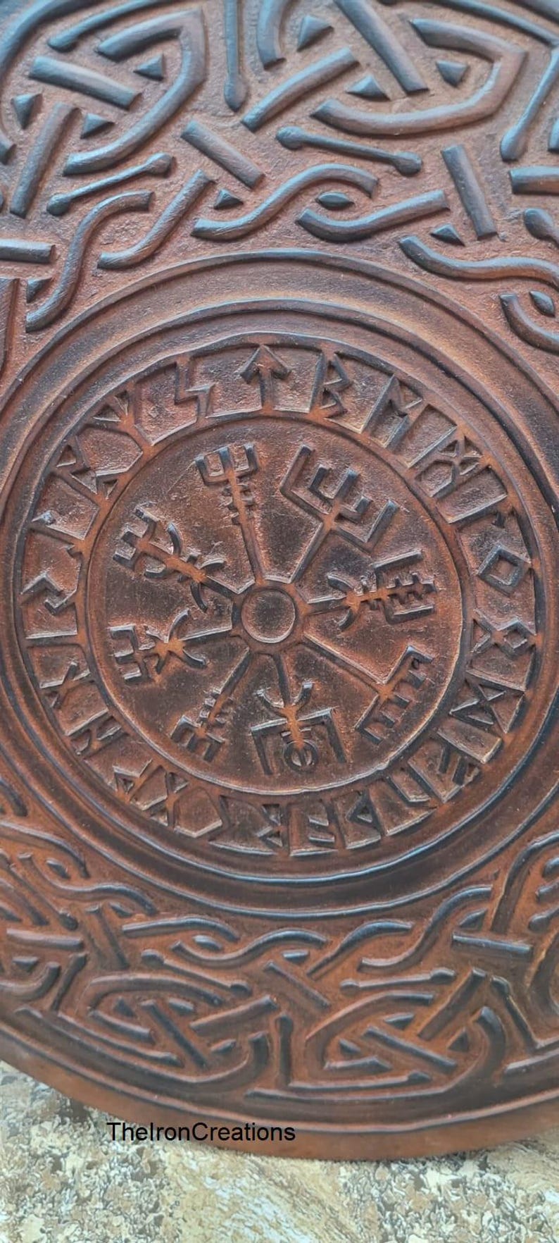 May include: Close-up of a brown, circular object with a raised, intricate design. The center features a compass-like symbol surrounded by runes. The outer edges display a repeating knot pattern. The text "TheIronCreations" is visible.
