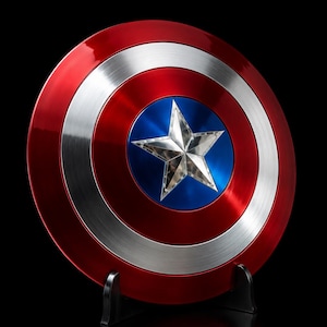 May include: A circular shield featuring a metallic finish with concentric rings of red and silver. The center consists of a blue circle containing a prominent, raised silver five-pointed star. The shield is supported by a black plastic display stand.