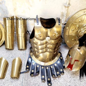 May include: A full set of antique brass Spartan armour, including a breastplate, shield, helmet, greaves, and arm guards. The armour is a shiny gold colour, with black leather accents. The shield features a lion's head.