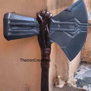 May include: A dark gray battle axe with a detailed design. The axe head has a rectangular hammer and a large, angled blade. The handle is dark brown with a wrapped grip. TheIronCreations is printed on the handle.