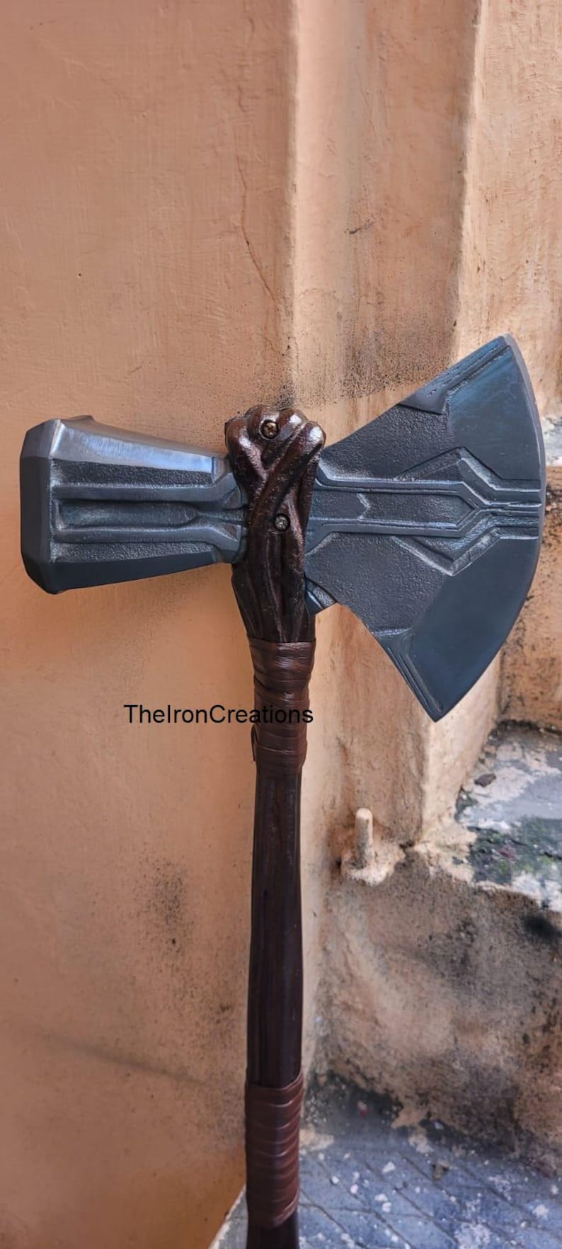 May include: A dark gray battle axe with a detailed design. The axe head has a rectangular hammer and a large, angled blade. The handle is dark brown with a wrapped grip. TheIronCreations is printed on the handle.
