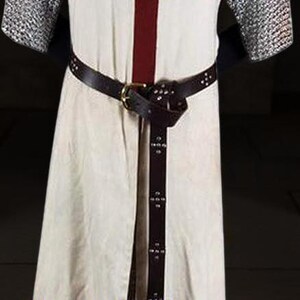 May include: Cream-colored medieval tunic with a large burgundy cross on the chest. The tunic features chainmail sleeves and a brown leather belt. A matching leather belt with decorative rivets cinches the waist. The text "TheIronCreations" is visible.