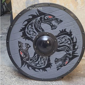 Könnte beinhalten: Ein runder, grauer Schild mit einem schwarzen Drachendesign und einem schwarzen Metallrand. Der Drache hat rote Akzente. Der Schild hat einen zentralen, schwarzen, abgerundeten Buckel und besteht aus Holzplanken. Dekorativer Artikel.
