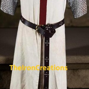 May include: Cream-colored medieval tunic with a large red cross on the chest. The tunic has chainmail sleeves and a black leather belt. The text "TheIronCreations" is visible on the bottom.