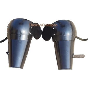 May include: Pair of black metal arm armor, likely for historical reenactment or costume use. Each piece features a rounded upper section and a cylindrical lower section, with leather straps for attachment. The armor is shown against a white background.