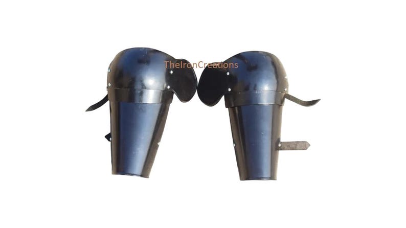 May include: Pair of black metal arm armor, likely for historical reenactment or costume use. Each piece features a rounded upper section and a cylindrical lower section, with leather straps for attachment. The armor is shown against a white background.
