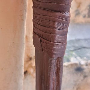 May include: Close-up of a dark brown wooden object with a wrapped leather detail. The leather is a lighter shade of brown and is wrapped around the wooden object. The text "TheIronCreations" is visible.