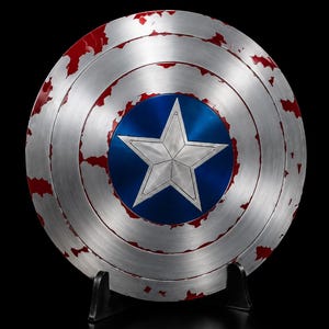May include: A circular metal shield featuring a brushed silver finish with distressed red paint accents. The center displays a vibrant blue circle with a large, five-pointed silver star. The shield is supported by a black plastic display stand.