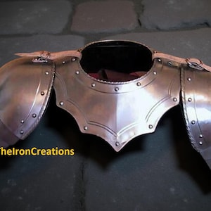 Può includere: Un'armatura per le spalle in metallo lucido con dettagli rivettati e cinghie in pelle. L'armatura ha una piastra pettorale centrale e protezioni per le spalle articolate. Il testo "ThelronCreations" è visibile in basso a sinistra.