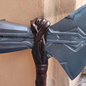 May include: A detailed, gray and brown prop axe with a textured handle and blade. The axe head is a dark gray color with etched lines. The handle is a dark brown color with a wrapped grip. The axe is a replica of a fantasy weapon.