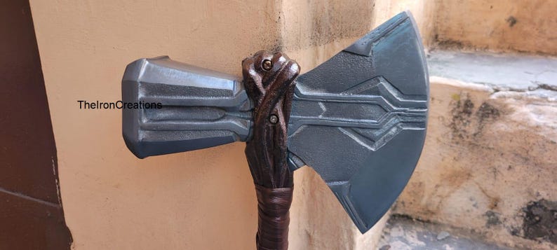May include: A detailed, gray and brown prop axe with a textured handle and blade. The axe head is a dark gray color with etched lines. The handle is a dark brown color with a wrapped grip. The axe is a replica of a fantasy weapon.