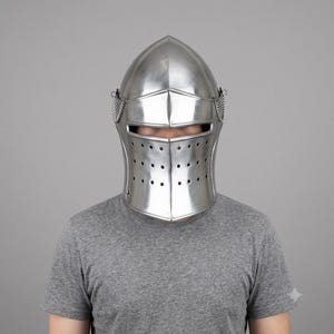 May include: A polished silver-colored metal knight helmet featuring a pointed top, a horizontal eye slit, and a lower section with multiple circular ventilation holes. Small silver chains are attached to both sides of the helmet near the temples.