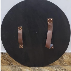 May include: A large, round, black shield with two brown leather straps secured by brass rivets. The shield is likely made of wood or a similar material, and the straps are for holding the shield.