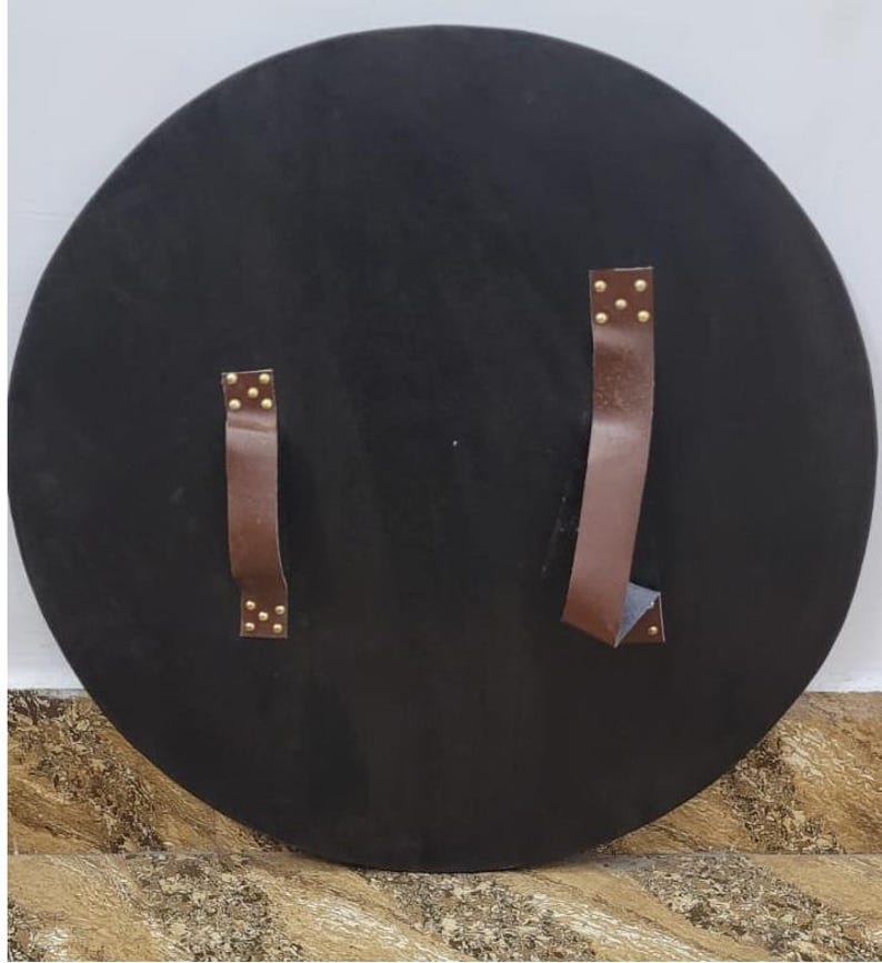 May include: A large, round, black shield with two brown leather straps secured by brass rivets. The shield is likely made of wood or a similar material, and the straps are for holding the shield.