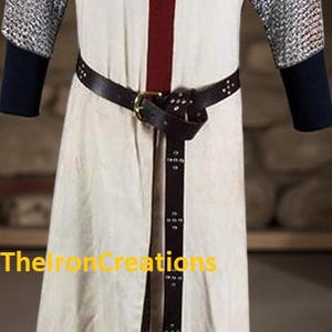 May include: Cream-colored medieval tunic with a large red cross on the front. The tunic has chainmail sleeves and a brown leather belt. The text "TheIronCreations" is visible on the lower left side.