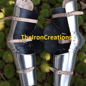 Könnte beinhalten: Paar mittelalterlicher Beinpanzer aus Stahl und Leder. Die Rüstung hat polierte Metallplatten, schwarze Lederteile und hellbraune Lederriemen mit Schnallen. Der Text "TheIronCreations" ist sichtbar.