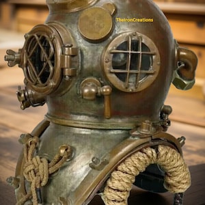 Op de afbeelding: Een antieke duikhelm met een verweerde bronzen afwerking. De helm heeft ronde kijkvensters, een bovenste ventilatieopening en diverse messing fittingen. Een dikke touwkraag en metalen steunen maken de vintage esthetiek compleet. De tekst "TheIronCreations" is zichtbaar.
