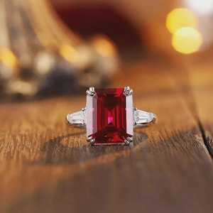 Puede incluir: Un anillo plateado con una gran gema rectangular de color rojo intenso como punto focal. Dos gemas blancas rectangulares más pequeñas flanquean la piedra principal. El anillo está sobre una superficie de madera, con luces borrosas de fondo.