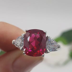 May include: A silver ring with a large, cushion-cut, ruby-red gemstone as the focal point. Two triangular, clear gemstones are set either side of the central stone. The background is soft and blurred.