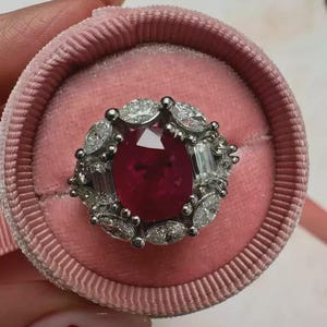 Pode incluir: Um anel luxuoso com uma grande pedra central de rubi de corte oval, rodeada por um halo de diamantes menores e diamantes baguette. O anel é cravejado em uma banda de prata ou ouro branco e apresentado em uma caixa de anel de veludo rosa.
