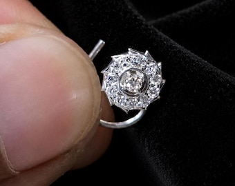 May include: A silver-toned nose ring featuring a circular floral-shaped setting. The center is adorned with a single round stone, surrounded by a ring of smaller, clear, sparkling stones. The metal band is thin and curved for piercing placement.