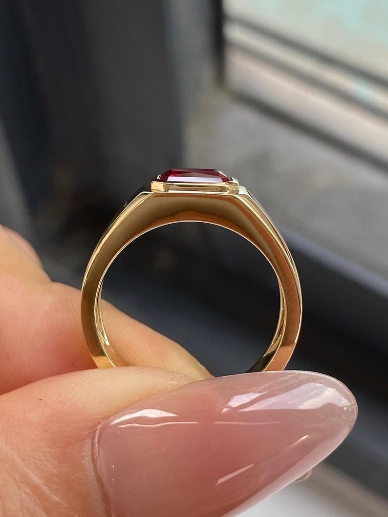 K&ouml;nnte beinhalten: Ein Goldring mit einem rechteckigen, tiefroten Edelstein in einer glatten, polierten Zargenfassung. Die Ringschiene besteht aus massivem Gold mit abgerundetem Profil und zeigt ein klares Design, das die leuchtende Farbe des Steins betont.