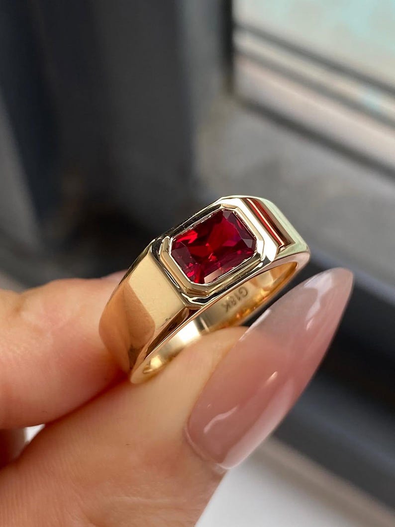 K&ouml;nnte beinhalten: Ein goldener Ring mit einem rechteckigen, tiefroten Edelstein in einer glatten, polierten Fassung. Das Band ist breit und massiv, wobei die Gravur G18K auf der Innenseite des Metalls deutlich sichtbar ist und den Goldgehalt des St&uuml;cks angibt.