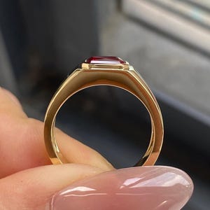 K&ouml;nnte beinhalten: Ein Goldring mit einem rechteckigen, tiefroten Edelstein in einer glatten, polierten Zargenfassung. Die Ringschiene besteht aus massivem Gold mit abgerundetem Profil und zeigt ein klares Design, das die leuchtende Farbe des Steins betont.