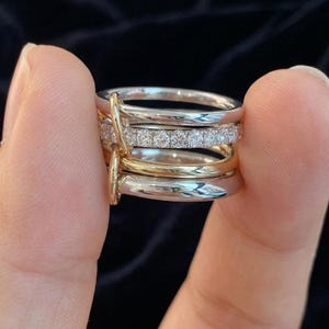 May include: A multi-band ring featuring a combination of silver-toned and gold-toned metal bands. One of the silver-toned bands is set with a row of small, clear, round-cut stones. A vertical metal loop connects the stacked bands together.