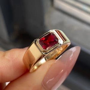 K&ouml;nnte beinhalten: Ein goldener Ring mit einem rechteckigen, tiefroten Edelstein in einer glatten, polierten Fassung. Das Band ist breit und massiv, wobei die Gravur G18K auf der Innenseite des Metalls deutlich sichtbar ist und den Goldgehalt des St&uuml;cks angibt.