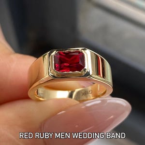K&ouml;nnte beinhalten: Ein goldener Ring mit einem rechteckigen roten Rubin in einer Zargenfassung. Das Band hat eine polierte Oberfl&auml;che und die Innenseite ist mit dem Text G18K graviert. Der Text RED RUBY MEN WEDDING BAND ist unten im Bild zu sehen.