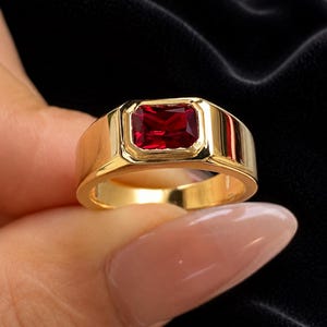 May include: A gold ring featuring a rectangular, faceted deep red gemstone set in a smooth, polished bezel setting. The band is wide and solid, crafted from a reflective gold-toned metal that contrasts with the vibrant, dark red stone centre.