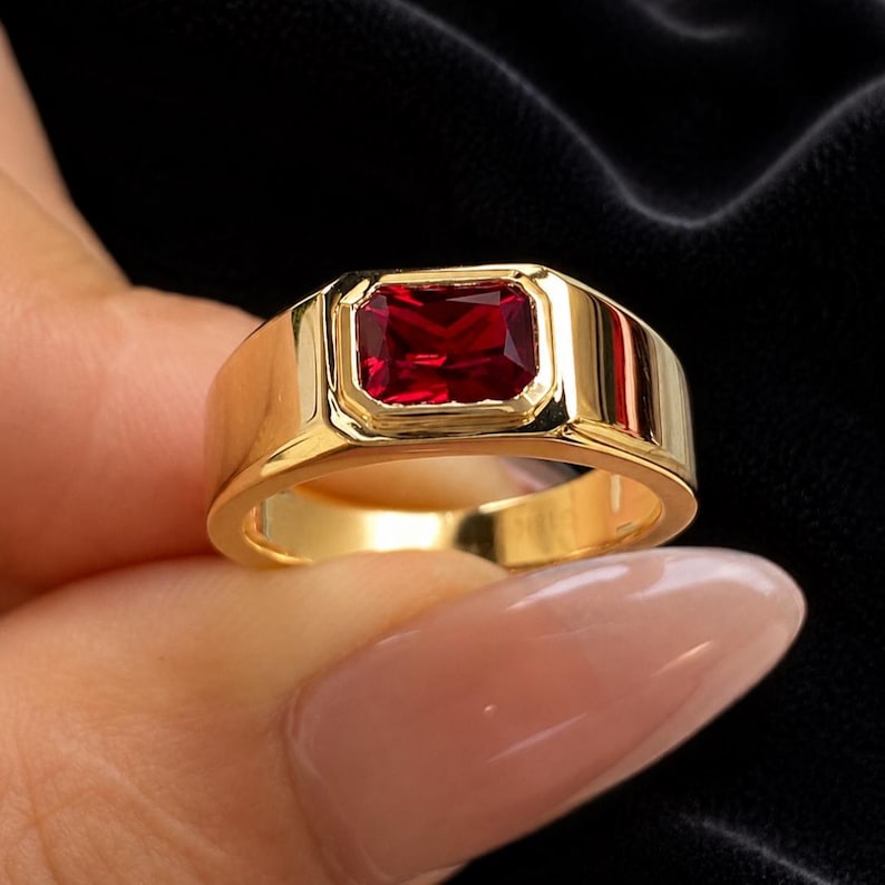 K&ouml;nnte beinhalten: Ein goldener Ring mit einem rechteckigen, facettierten tiefroten Edelstein in einer glatten, polierten Zargenfassung. Die Ringschiene ist breit und massiv und besteht aus reflektierendem goldfarbenem Metall, das einen Kontrast zum Stein bildet.