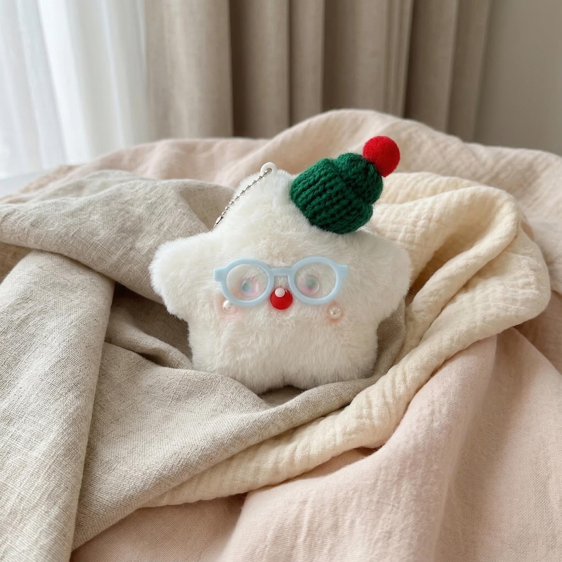 May include: A white plush star-shaped keychain with a green knitted hat and a red pom-pom. It has light blue glasses, a red nose, and small pink pearls. The keychain is on a light-colored fabric.