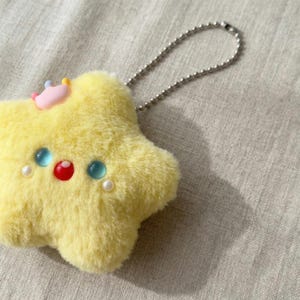 May include: A yellow plush star-shaped keychain with a small pink crown and decorative blue, red, and white embellishments. The keychain has a silver ball chain attached to the top. The plush toy has a soft, fuzzy texture.