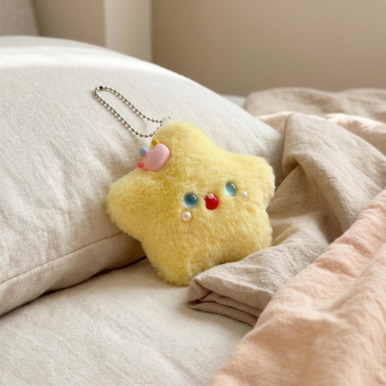 May include: A yellow star-shaped plush keychain with a smiling face, blue and red accents, and a pink crown. The keychain has a silver chain and is resting on a bed with neutral-colored linens.