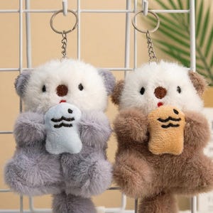May include: Two plush bear keychains. One is gray and white, the other is brown and white. Each bear holds a small plush item. The keychains have silver metal rings and chains.